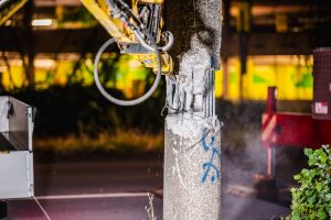 Ein hydraulischer Greifarm mit Abbruchwerkzeug zerbricht bei Nacht einen Betonmast. Sichtbare Stahlbewehrung ragt aus dem beschädigten Bereich heraus, während Staub und Trümmer die Szene umgeben. Der Mast trägt blaue Markierungen, vermutlich zur Kennzeichnung. Im Hintergrund sind eine Straße und ein beleuchtetes Gebäude zu sehen. Die Szene ist durch künstliches Licht erhellt und zeigt aktive Infrastrukturarbeiten.
