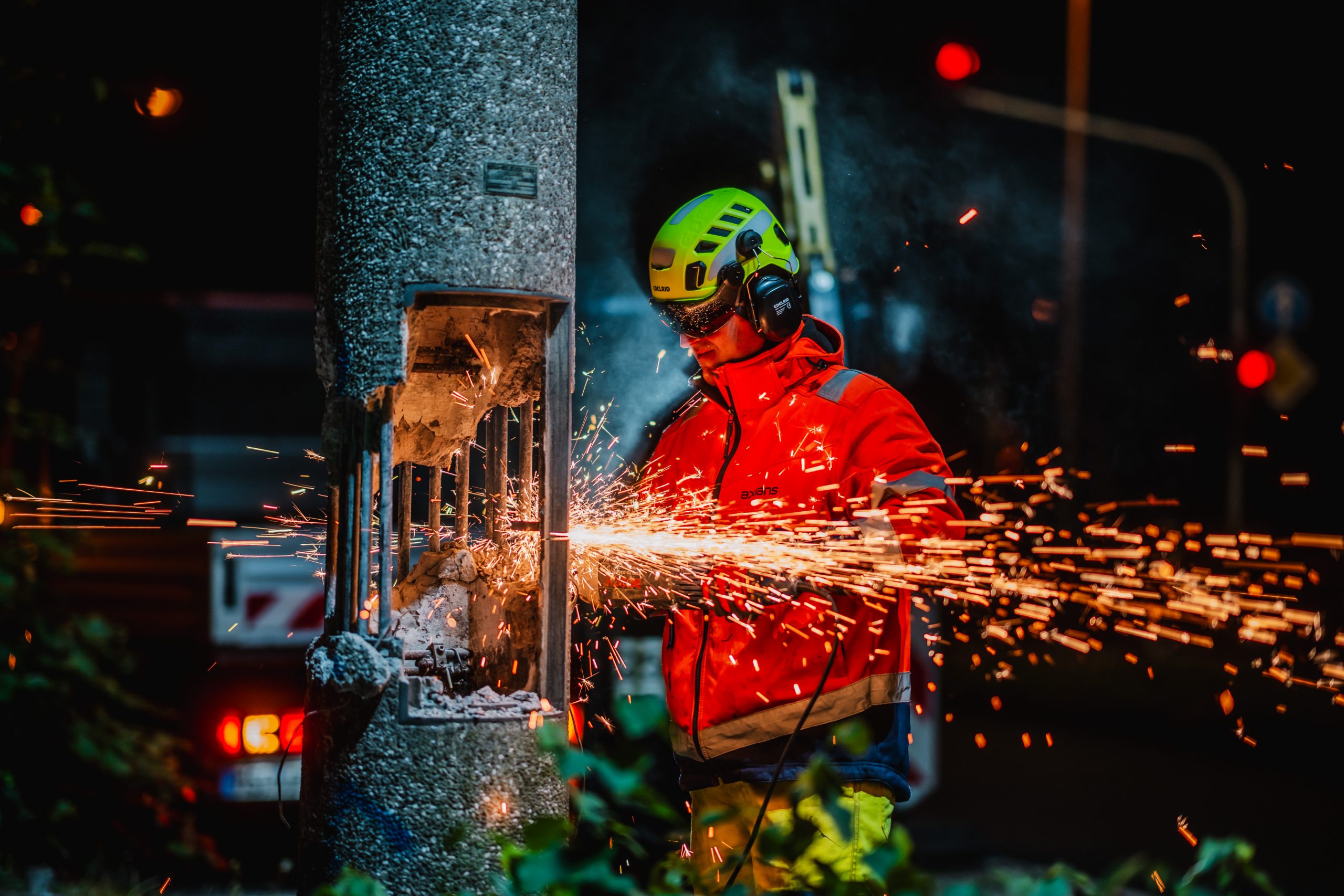 Eine Person in Schutzkleidung – Helm, Gehörschutz und Warnweste – schneidet nachts mit einem Winkelschleifer einen Betonmast. Funken sprühen hell auf, während Metall freigelegt wird. Im Hintergrund sind Straßenbeleuchtung und Fahrzeuglichter sichtbar, die die Szene dramatisch ausleuchten. Die Situation wirkt wie eine Wartungs- oder Abrissarbeit.
