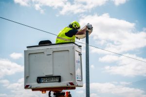 Ein Arbeiter mit Helm und Warnweste steht in einem weißen Arbeitskorb, der an einem hydraulischen Hubarm befestigt ist. Er installiert oder wartet eine Überwachungskamera an einem hohen Metallmast. Der Himmel ist teilweise bewölkt, Stromleitungen verlaufen horizontal im Hintergrund. Die Szene zeigt Infrastrukturarbeit im öffentlichen Raum